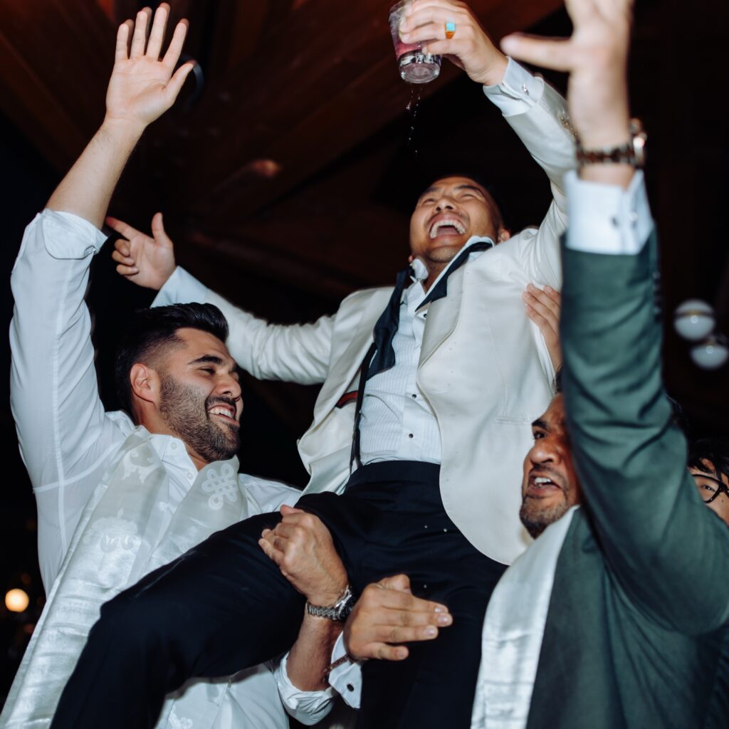 Groom being held up by friends, smiling, and dancing with a drink