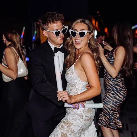 Bride and groom smiling with heart glasses dancing in a dark, crowded room